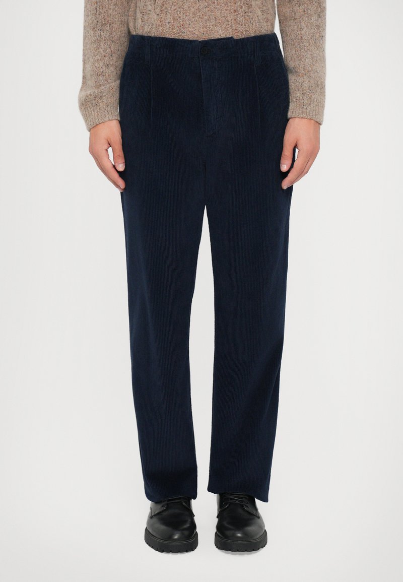Persona vestida con pantalones de pana azul marino, zapatos de cuero negros y un suéter de punto beige, de pie contra un fondo blanco liso.