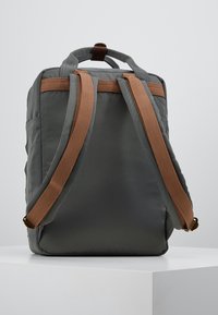 Sac à dos en tissu gris avec des sangles ajustables marron et une poignée supérieure. Texture lisse, forme rectangulaire et détails de design minimalistes.