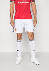 Camisa de futebol vermelha com detalhes pretos e logotipo, combinada com calções brancos com emblema do clube. Meias brancas com riscas pretas e chuteiras.