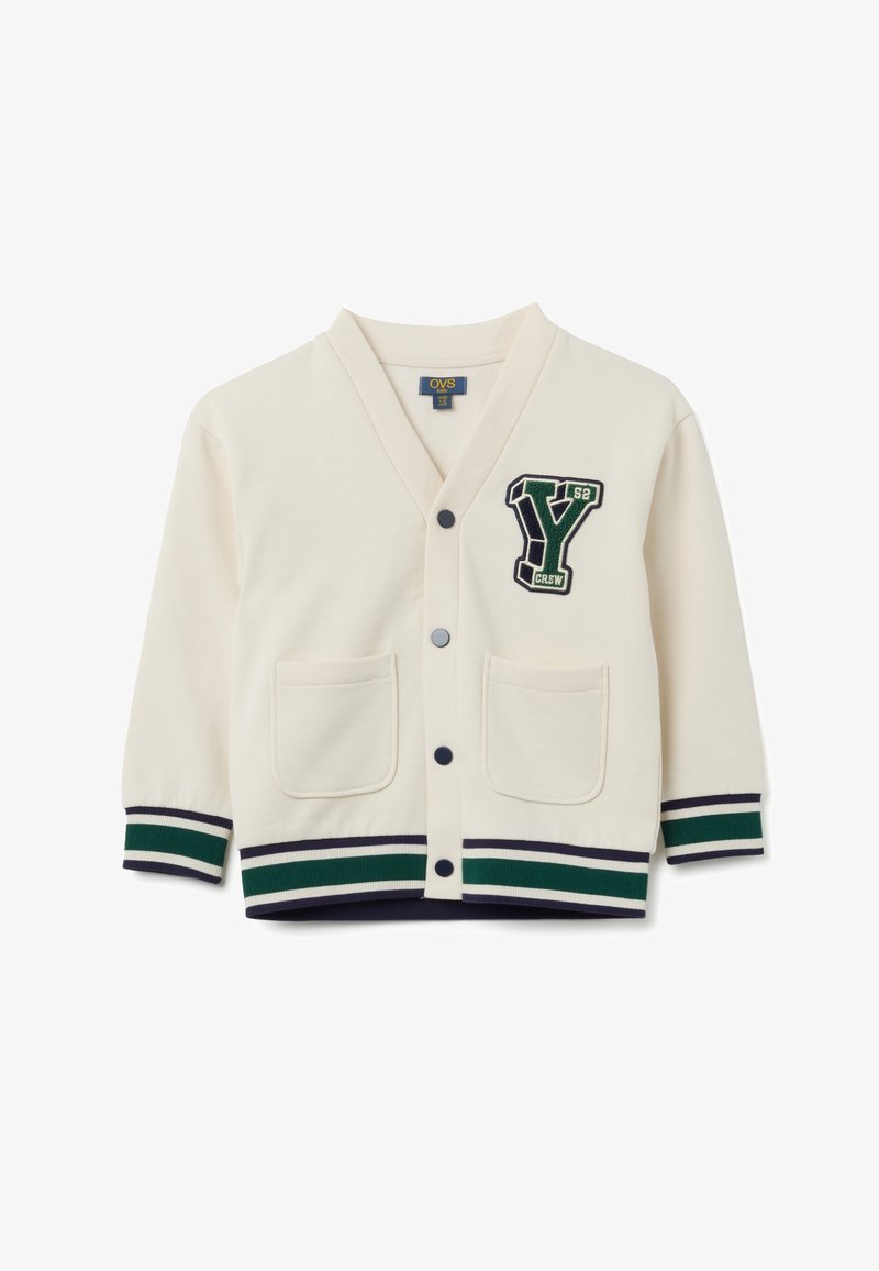 Cárdigan crema para niños con botones negros, dos bolsillos frontales, puños y dobladillo a rayas verdes y negras, y un parche en estilo universitario con la 'Y' verde en el pecho.