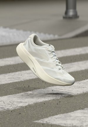 Bílá sportovní bota ve vzduchu nad přechodem pro chodce na šedé asfaltové silnici se drobnými úlomky po ní.