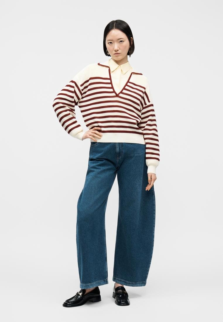 Femme portant un pull à rayures crème et bordeaux à col en V, une chemise à col crème, un jean bleu large et des mocassins noirs, debout avec une main sur la hanche.