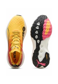 Zapatillas deportivas amarillas y rojas con parte superior texturizada, suela de patrón negro y rosa, y cordones amarillos; se puede ver la marca.