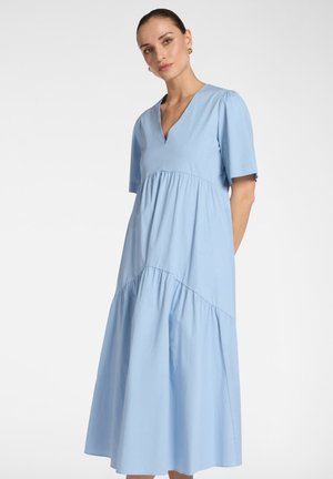 Donna che indossa un abito midi a balze azzurro chiaro con scollo a V, maniche corte e orecchini a cerchio dorati su uno sfondo neutro.