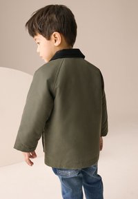 Veste vert olive avec un col noir, présentant une coupe décontractée et des manches longues. Portée avec un jean bleu, elle affiche un design simple et une texture lisse.