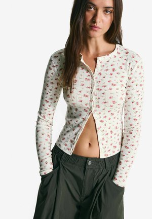 Woman with long dark hair wearing a white floral buttoned cardigan partially open, and dark loose-fitting pants with hands in pockets.
