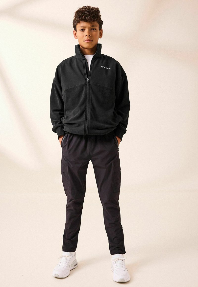 Schwarze Fleece-Zipjacke mit kontrastierendem Stoffeinsatz, kombiniert mit schwarzen Cargo-Hosen und weißen Sneakers, vor einem neutralen Hintergrund.