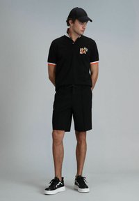 Zwarte polo met oranje en witte accenten, met een ontwerp van een borstzak. Zwarte shorts en zwarte sneakers met witte zolen maken de look compleet.