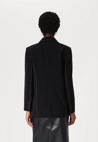 Blazer nero su misura con maniche lunghe, cuciture gialle a contrasto, colletto a punta e polsini abbottonati. Tessuto liscio con un design strutturato.