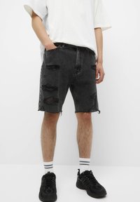 Shorts en denim noir avec un ourlet effiloché et plusieurs déchirures sur le devant. Portés avec un t-shirt blanc ample et des baskets noires avec des rayures blanches.