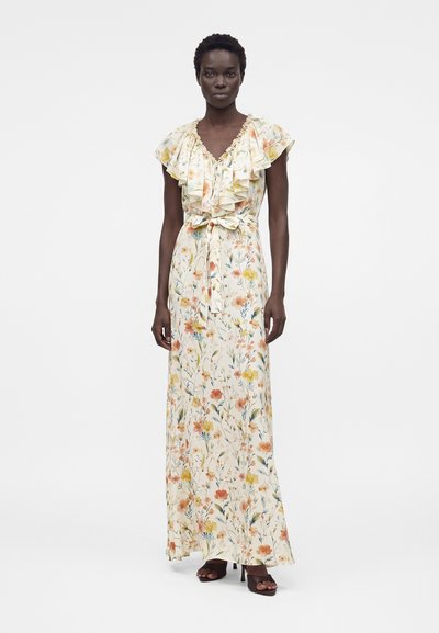 Γυναίκα που φοράει ένα μακρύ, κρεμ φόρεμα με λουλούδια, με πλισέ μανίκια και ζώνη στη μέση, σε συνδυασμό με σκούρα πέδιλα ανοιχτής μύτης.