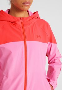 Różowa i czerwona kurtka rozpinana z kapturem, o gładkiej teksturze, z logo 'Under Armour' na klatce piersiowej. Kontrastowe bloki kolorów.