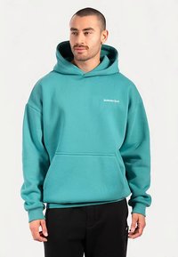 Tealfärgad huvtröja i bomullsblandning, med kenguruficka och ribbstickade muddar. Litet vitt logo på övre vänstra bröstet.