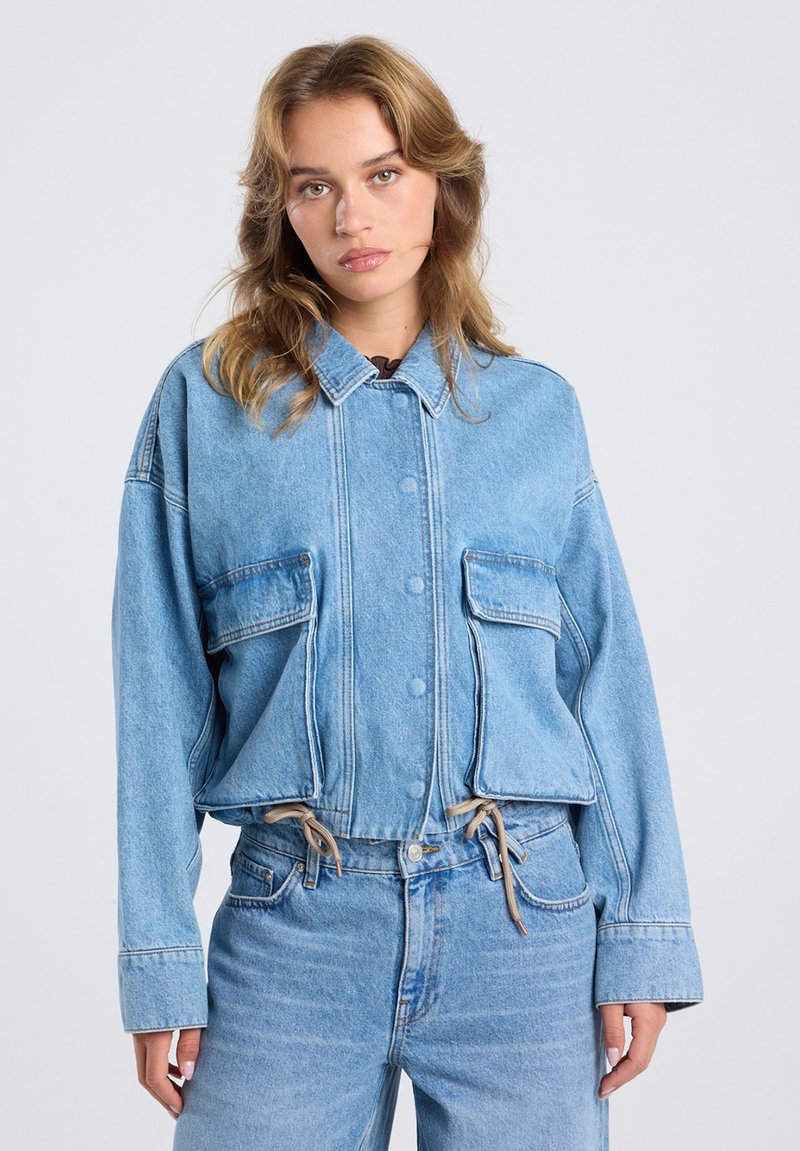 Jonge vrouw met lichtbruin haar, gekleed in een lichtblauwe denimjas met grote voorkruiken en bijpassende blauwe jeans tegen een effen achtergrond.