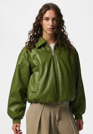 Donna con capelli ricci che indossa una giacca di pelle verde con cerniera e pantaloni beige plissettati su sfondo bianco semplice.