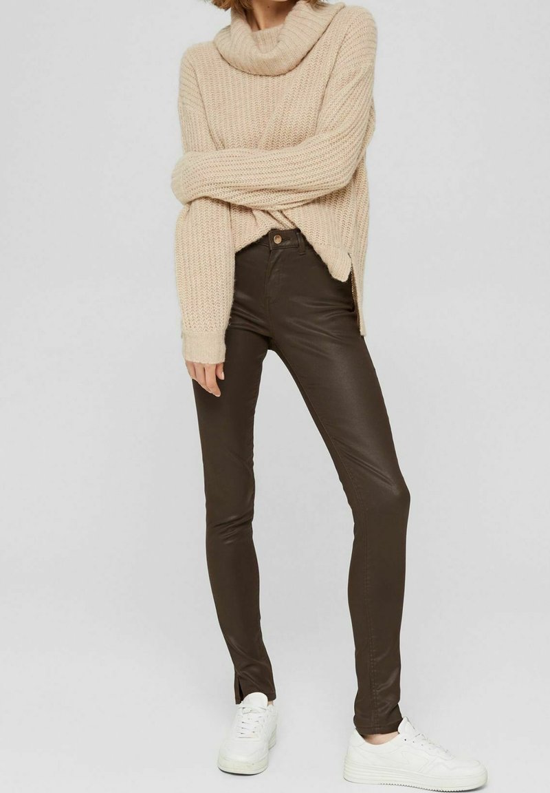 Beige ribbad stickad tröja med en cowl-hals, bärs över mörkbruna, åtsittande leggings och vita sneakers, vilket visar en avslappnad lager-stil.