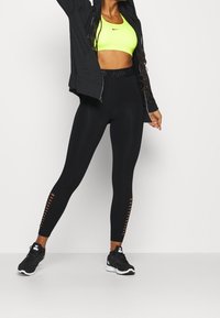 Žena v neónově žlutém sportovním brá, černé zipové mikině, černých legínách s bočními výřezy a černých teniskách Nike stojící proti bílé zdi.