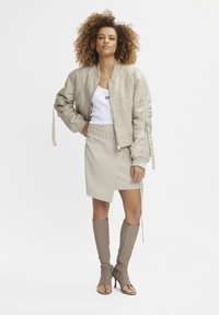 Beige bomber jacket with textured fabric, fitted tank top, asymmetric beige skirt with studs, and taupe knee-high split-heel boots.