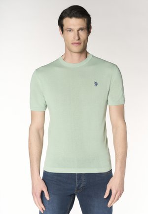 Uomo con maglietta a maniche corte in maglia verde chiaro e jeans blu, in piedi con espressione neutra su sfondo semplice.