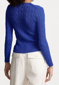 Blå stickad tröja med kabelmönster, figursydd midja och ribbade ärmslut. Buren med ljusa byxor med hög midja och bakfickor.