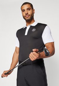 Čierno-bielý golfový polo trikoty s krátkymi rukávmi, golierom a kruhovým logom na hrudi. Model drží golfovú hokejku a má na sebe čierne nohavice.