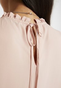 Rückenansicht einer Person, die eine hellrosa Bluse mit Rüschenausschnitt und gebundenem Stoffband trägt, sowie eine silberne Halskette.