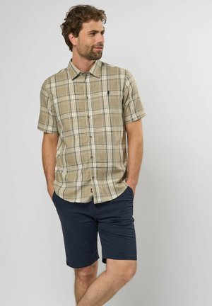 Camisa de cuadro beige de manga corta con detalles en verde y blanco, cuello abotonado y logo en navy. Combinada con pantalones cortos navy.