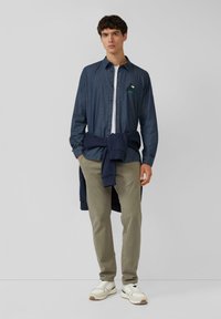 Ciemnoniebieska koszula na guziki z krótkimi rękawami, haftowany wzór żółwia na piersi, beżowe spodnie i białe trampki z szarymi akcentami.