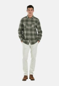 Camicia a quadri verdi con bottoni e due tasche sul petto, abbinata a pantaloni bianchi e scarpe stringate marroni. Tessuto morbido, vestibilità su misura.