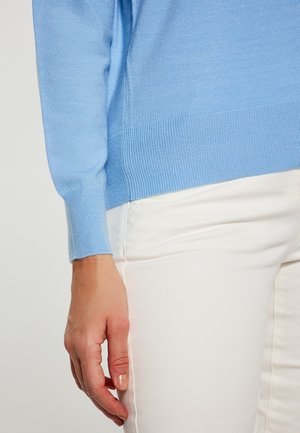 Hellblauer Strickpullover mit gerippten Bündchen, getragen über weißen Hosen. Das Material wirkt weich und leicht, mit einer glatten Textur.