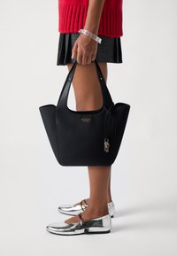 Sac en cuir noir à texture grainée avec de courtes poignées, logo doré et accent de porte-clés en argent. Modèle portant des chaussures plates argentées, debout.