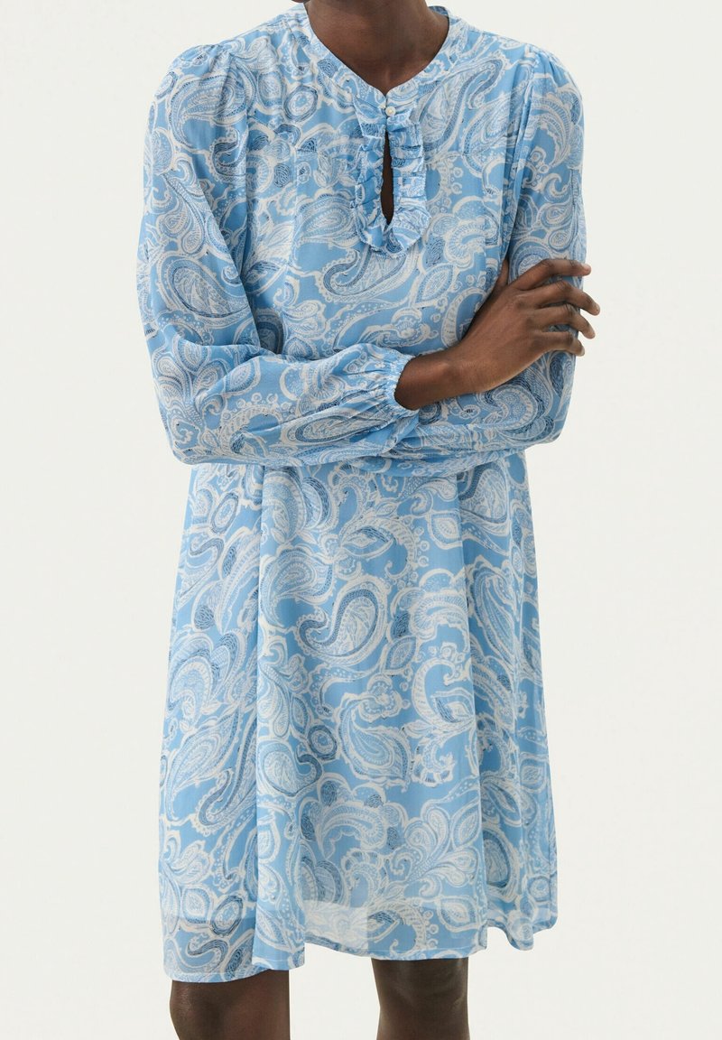 Personne portant une robe bleue avec des motifs paisley blancs, des manches longues et un col en trou de serrure à volants, debout, les bras croisés.