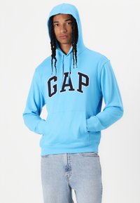 Light blue hoodie with a large "GAP" logo in navy. Features a kangaroo pocket, drawstring hood, and ribbed cuffs. Worn with light denim jeans.