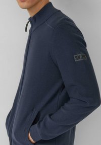 Marineblauwe gebreide ritssluiting vest met een hoge kraag, geribbelde manchetten en zijzakken. Heeft een logopatch op de linkerarm.