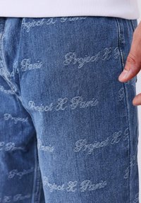 Jeans de denim azul con un patrón de texto tonal "Project H Paris" en toda su superficie, en un bordado ligeramente elevado. Corte recto clásico.