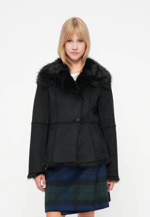 Veste noire avec col en fausse fourrure, présentant un design ajusté et une fermeture à boutons. Portée avec une jupe à carreaux dans des tons de bleu et de vert.