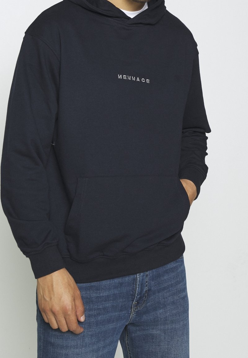 Homme portant un sweat à capuche noir avec l'inscription « MENNACE », jean bleu, main dans la poche avant, sur un fond clair uni.