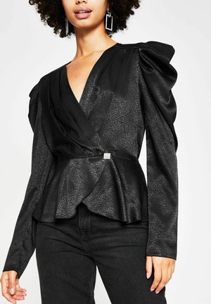 Mujer con blusa negra cruzada con hombros abullonados, textura sutil de estampado animal, hebilla de pedrería, combinada con jeans negros y pendientes geométricos.