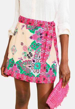 Vrouw draagt een witte blouse en een kleurrijke bloemen mini-rok met een roze riem, houdend een roze gestructureerde clutchtas vast.