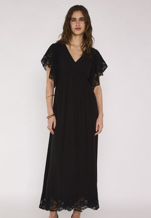 Femme portant une longue robe noire avec un bord en dentelle sur les manches courtes et l'ourlet, debout devant un fond clair uni, face à l'avant.