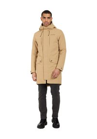 Tandy parkas med huva, frontdragkedja och tryckknappar; har två framfickor och rak skärning. Bärs med mörka jeans och svarta stövlar.