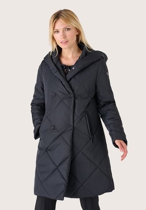 Donna bionda che indossa un lungo cappotto trapuntato nero con ampio colletto, dettagli con bottoni e cerniere, in piedi davanti a uno sfondo semplice.