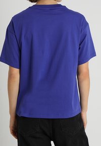 Osoba ubrana w gładką niebieską koszulkę z krótkim rękawem i czarne spodnie, pokazana od tyłu na jednolitym tle.