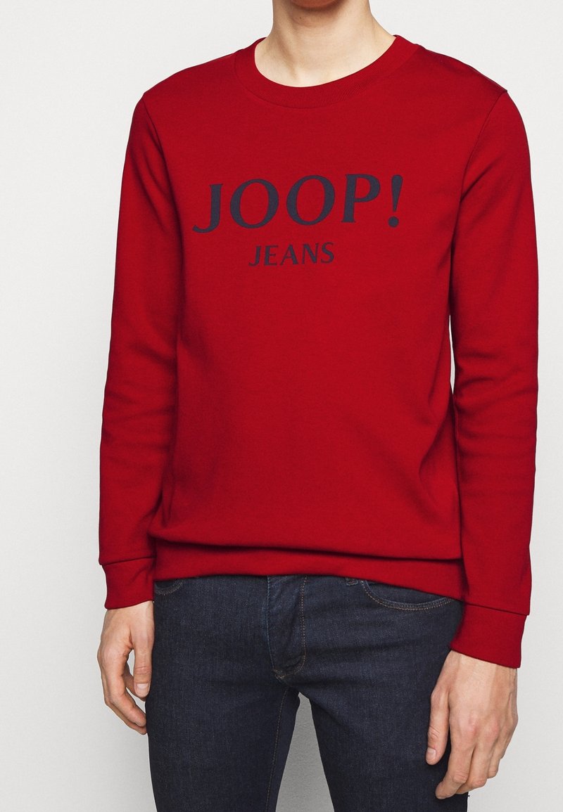 Personne portant un t-shirt à manches longues rouge avec le texte "JOOP! JEANS" sur le devant et un jean bleu foncé, debout devant un fond uni.