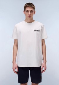 Beige katoenen T-shirt met korte mouwen, met een zwart logo op het linkerborstgebied. Gecombineerd met donkerblauwe shorts.