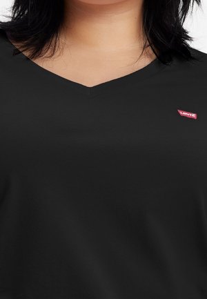 Mujer con una camiseta negra de cuello en V con una pequeña etiqueta roja del logo de Levi's en el pecho izquierdo, mostrando cuello y torso superior.