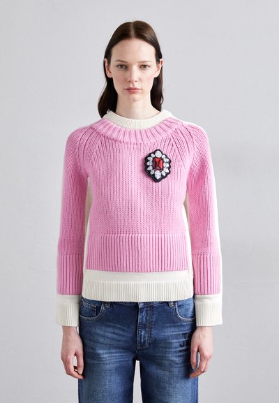 Pull en tricot rose avec texture côtelée, accents blancs, col rond et une patch noire avec un design décoratif sur la poitrine.