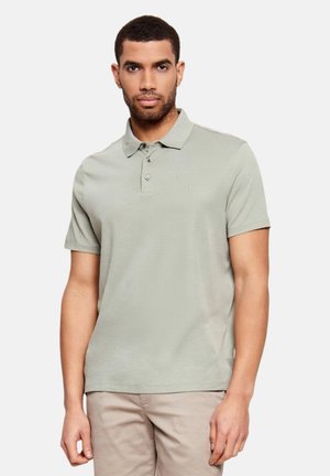 Hombre con camisa polo de manga corta color gris claro y pantalones beige, de pie contra un fondo claro y liso, mirando hacia adelante.