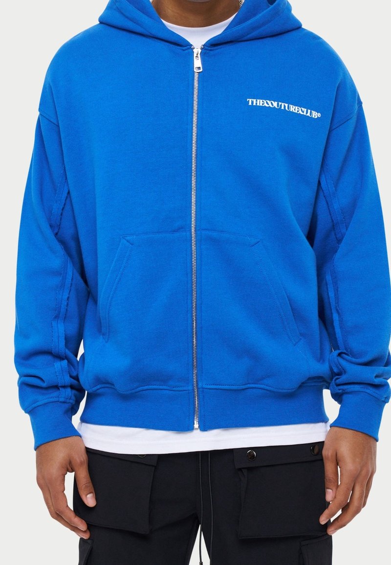 Niebieska bluza z zamkiem, wykonana z miękkiego materiału, z przednią kieszenią, kapturem oraz napisem "THE COUTURE CLUB" nadrukowanym na lewej piersi.