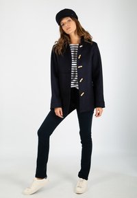 Mörkblå ullrock med träknappar, kombinerad med en randig skjorta, svarta byxor och vita sneakers. Modell som bär en matchande keps.